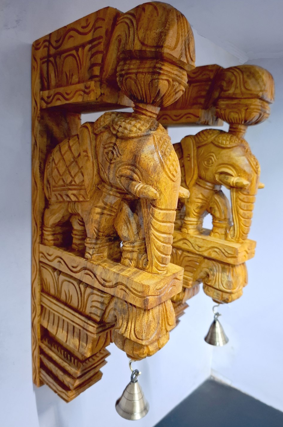A pair of wooden elephant wall corbels by JLine Arts, featuring a vibrant teak color finish and hanging brass bells. These hand-carved wall brackets are perfect for door entrance wall decor or as unique wall hangings