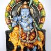 The 15 Krishna Wall Hanging features a blue figure playing a flute while seated on a yellow cow, set against an ornate backdrop with an elegant arch, making it perfect for living rooms, gardens, or temples.