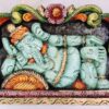 A wooden Ganesha wall panel features the deity reclining elegantly with intricate details and a crown, set against a floral arch. This Ganesh Statue combines traditional charm with exquisite craftsmanship, epitomizing artistry in its design.