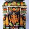 A vibrant Wooden Ganesh Statue, featuring the deity Ganesha with four arms seated on a lotus, is surrounded by ornate pillars and decorative elements. This Ganesha Wall Panel captures divine elegance and craftsmanship in every detail.